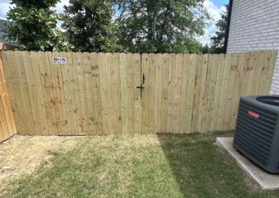 A wooden privacy fence with a closed gate and a small sign reading Beware of Dog, next to a white brick house and an outdoor air conditioning unit, with grass and trees in the background.