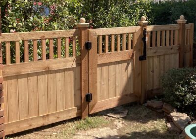 A wooden garden gate with vertical slats and decorative posts is surrounded by bushes and greenery, with a stone pathway leading up to it. Sunlight casts shadows across the gate and yard.