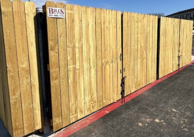 A tall wooden privacy fence with a gate stands beside a blacktop area marked with a red curb. A sign on the fence displays Bills and a phone number. The sky is clear and blue.