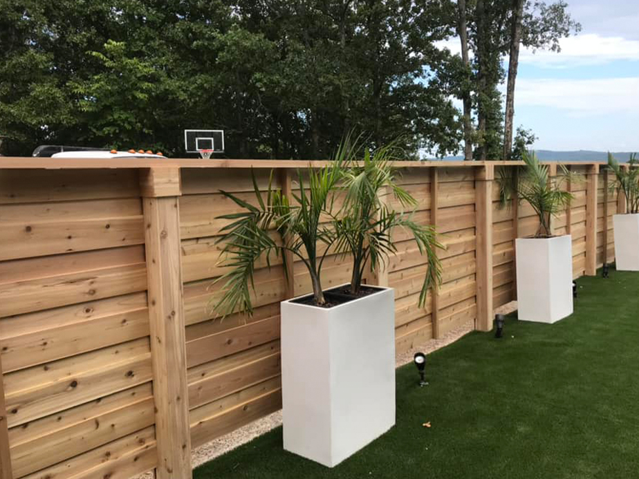 Tall wooden fence with rectangular white planters holding palm plants lined up along artificial grass. Small spotlights are placed at the base of the fence. Trees and a basketball hoop are visible in the background.