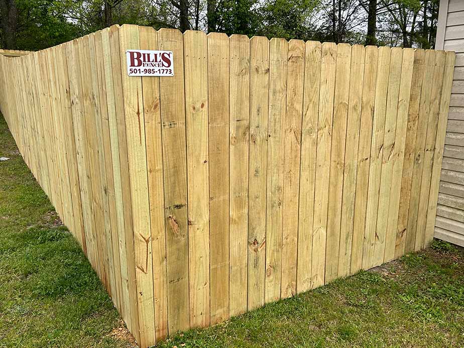 A tall wooden privacy fence encloses a yard, with a sign reading BILLS Fence 501-985-1773 attached to one side. The fence stands on grass with trees and a building visible in the background.