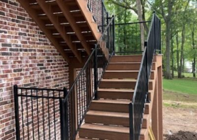 A wooden staircase with black metal railings is attached to a brick building. The stairs lead to an upper level, and there are trees and grass visible in the background.