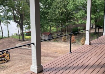 View from a covered wooden deck overlooking a dirt yard with trees, a small shed, and construction equipment. A lake is visible in the background beyond the trees. Black railing lines the edge of the deck.