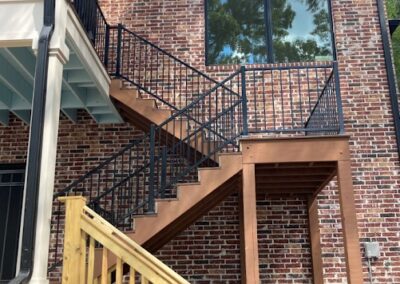 A two-story brick building with a wooden staircase leading up to a black metal staircase that continues to a second-floor entrance, beside a large window reflecting trees and sky.