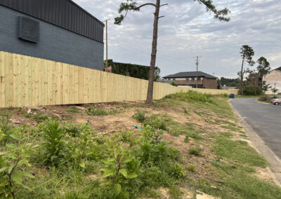A newly installed wooden fence runs along the edge of a dry, patchy, grassy area beside a paved road, with a gray building and a few trees in the background under a cloudy sky.
