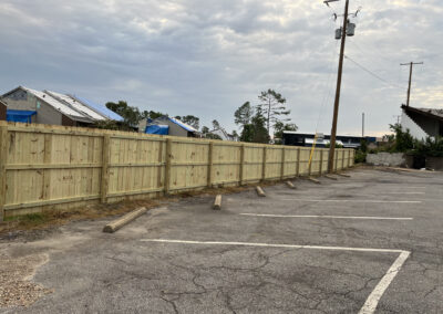 Empty parking lot with faded lines and parking stops, bordered by a newly built wooden fence. Houses with tarped roofs are visible in the background under a cloudy sky.