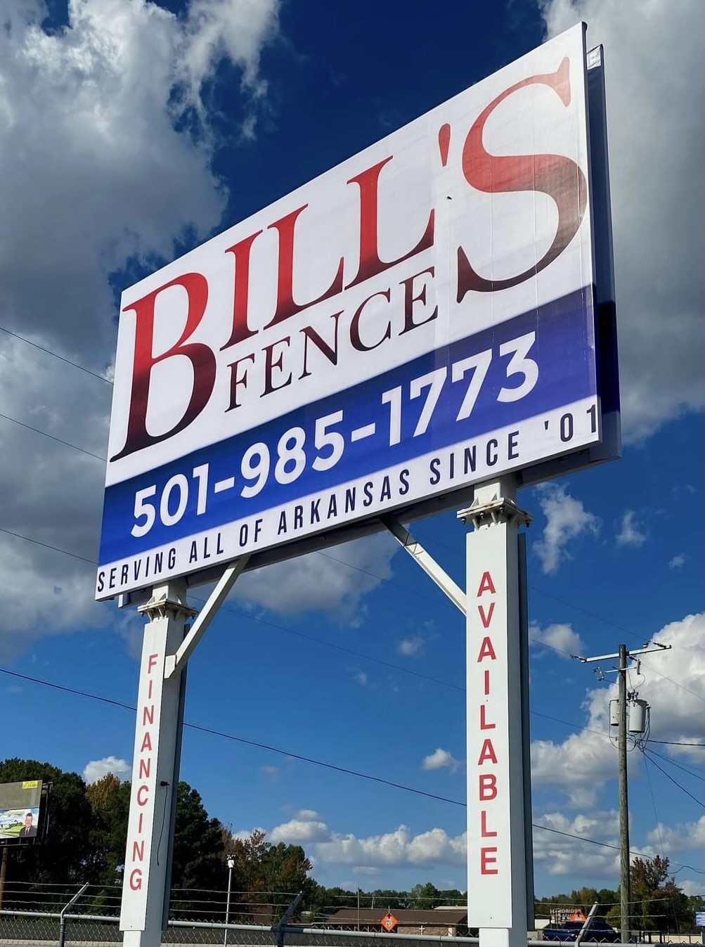 A large roadside sign for Bills Fence with a phone number, 501-985-1773. Text also reads, Serving all of Arkansas since 01 and Financing Available on side panels. The sky is partly cloudy in the background.