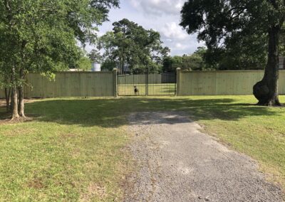 A paved driveway leads to a wooden fence with a black metal gate. Beyond the gate, a dog stands on green grass. Trees frame the scene, and the sky is partly cloudy.