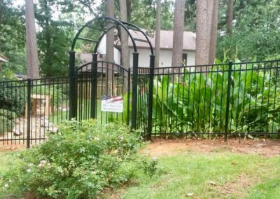 A black metal fence with an arched gate encloses a garden with tall green plants and bushes, set in a grassy yard with trees and a house in the background.