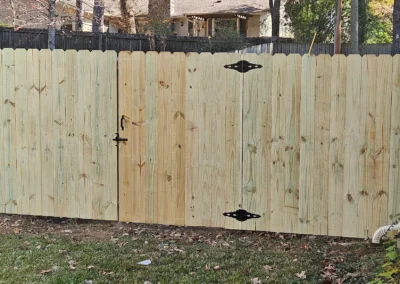 A new wooden fence with a double gate and black metal hinges stands between a brick house and an older gray fence. Fallen leaves are scattered on the grass in front.
