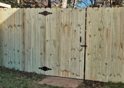 A wooden privacy fence with a gate is secured by black metal hinges and a latch, standing next to a brick wall in a grassy backyard area with trees in the background.