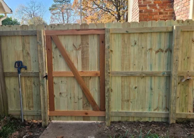 A newly built wooden fence with a gate stands between two brick buildings. The fence is made of light-colored wood and has a diagonal support on the gate. A shovel leans against the left side of the fence.