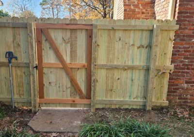 A newly built wooden fence with a gate stands between a brick wall and an older wooden fence. A garden hose holder is mounted on the left, and fallen leaves are scattered on the ground.