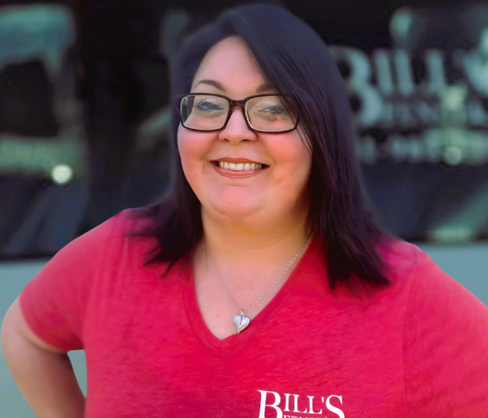 Sammie Conelly with dark hair and glasses smiles at the camera. She is wearing a red V-neck shirt with BILL’S written on it and a heart-shaped necklace. The background is blurred with indistinct signage.