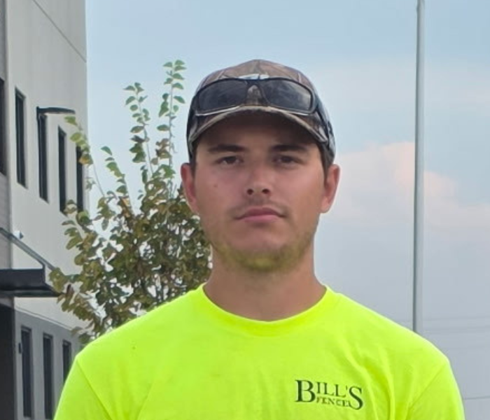 A man wearing a neon yellow BILLS FENCES T-shirt, camouflage cap, and sunglasses on his hat stands outdoors in front of a building and a small tree.