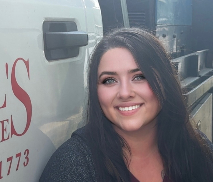 A woman with long dark hair and blue eyes is smiling while sitting next to a white vehicle with red lettering. She is wearing a dark top, and sunlight is shining on her face.