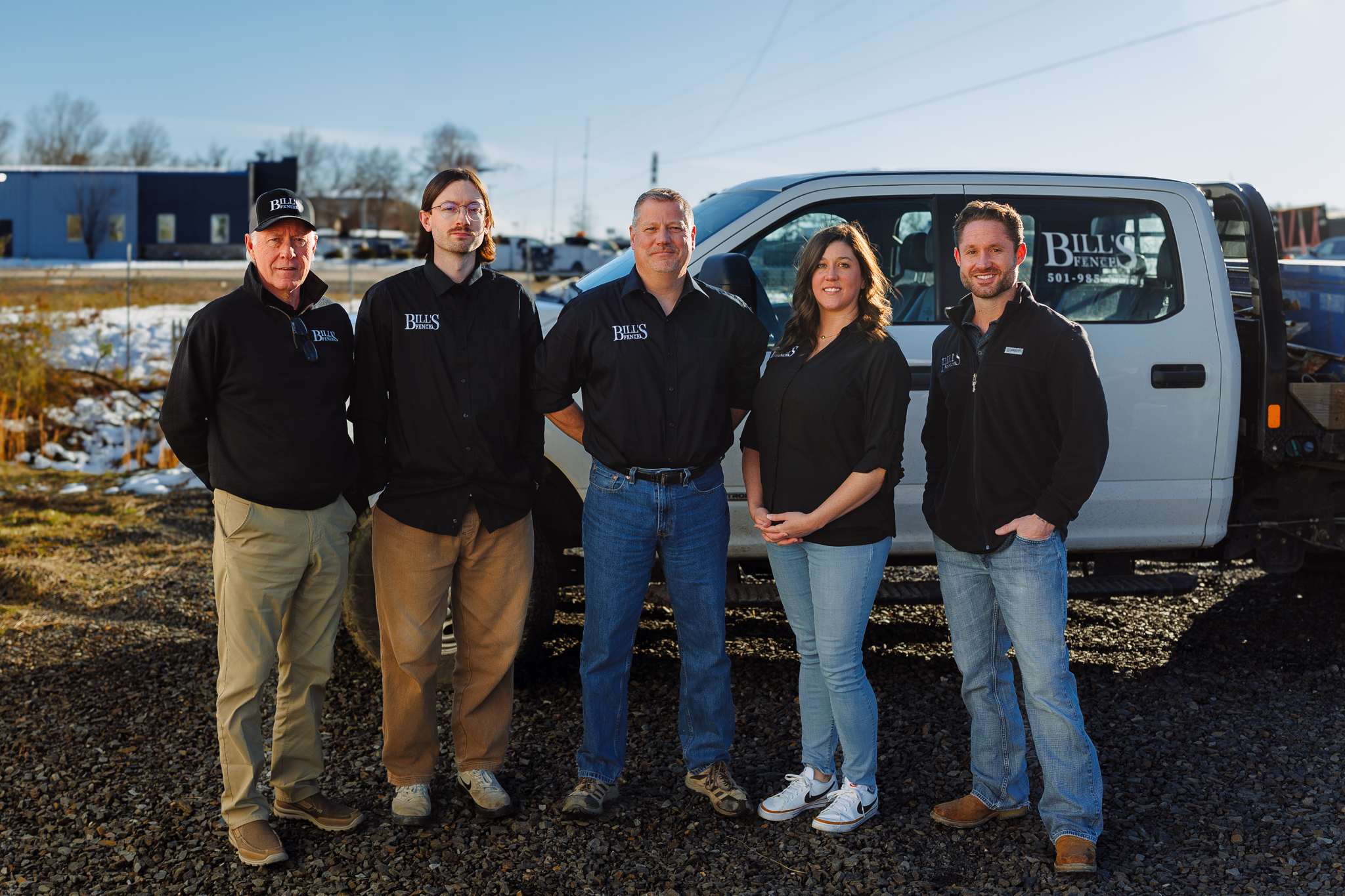 Five people stand side by side in front of a pickup truck on a gravel lot. They are casually dressed, mostly in black shirts, and are posing and smiling for the camera. The truck has a company logo on its door.