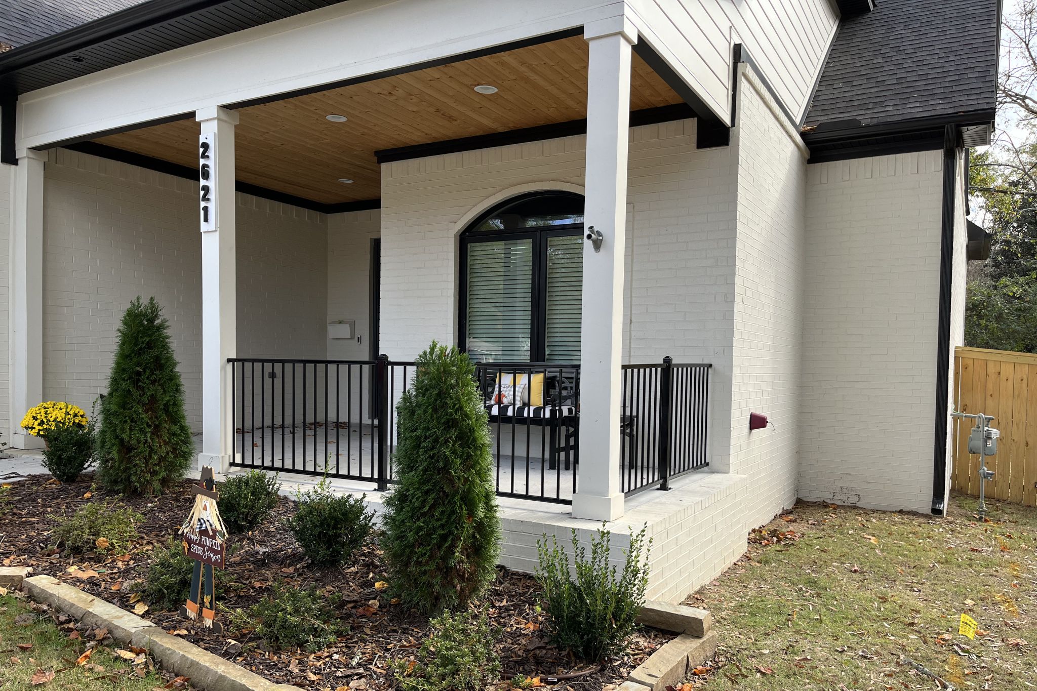A modern house with white brick exterior, a small porch with black railing, potted shrubs, and fall decorations including a scarecrow figure and yellow flowers in the front yard. The house number 2621 is on a porch column.