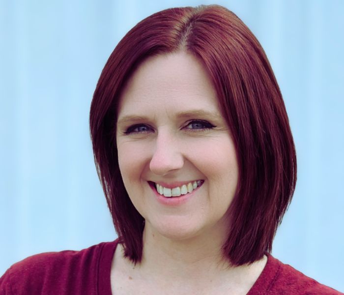 A woman with straight, shoulder-length reddish-brown hair smiles at the camera. She is wearing a maroon top and is standing in front of a light blue background.