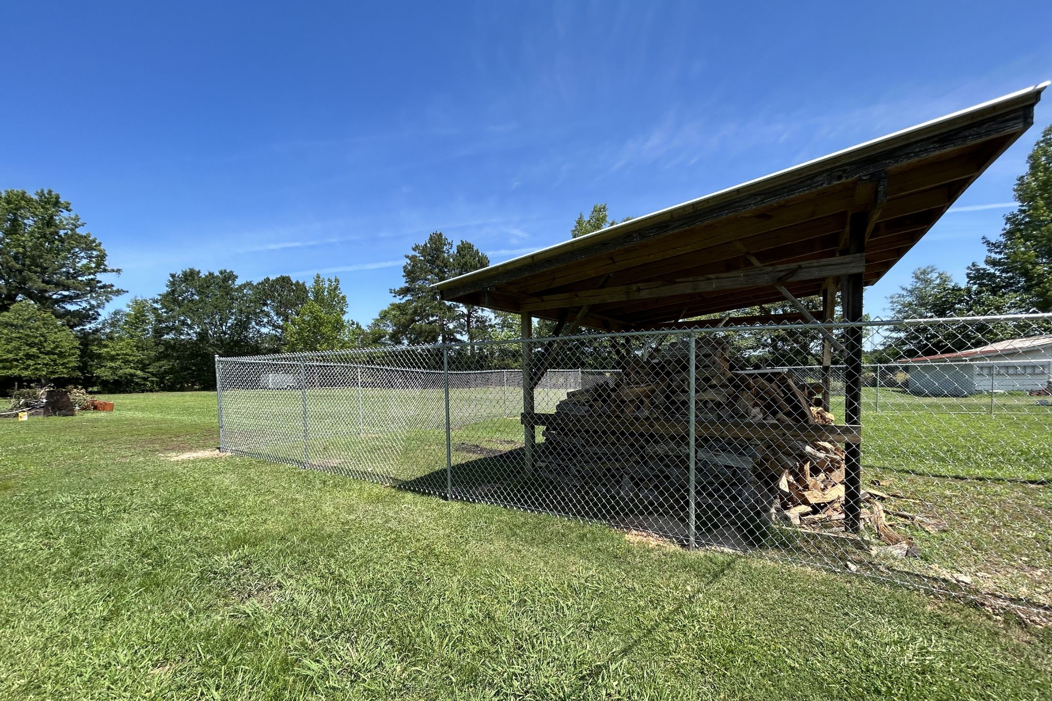 A stack of firewood is stored under a wooden shelter with a metal roof, enclosed by a chain-link fence, in a grassy yard with trees and buildings in the background under a clear blue sky.