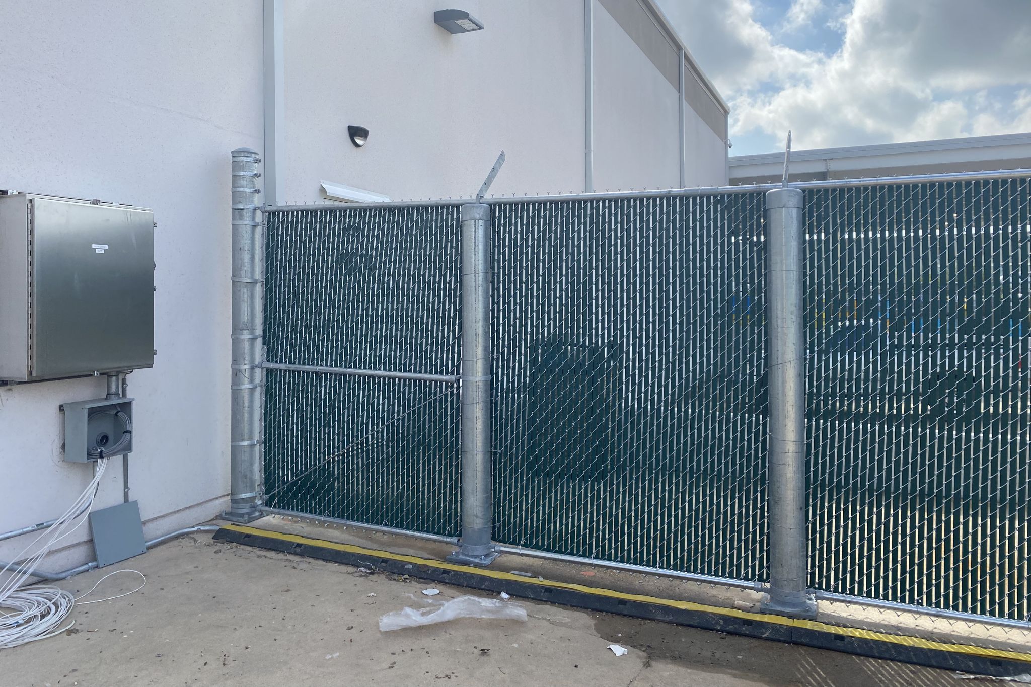 A tall metal chain-link fence with privacy slats borders a concrete area next to a white building. Electrical boxes and cables are mounted on the wall, and the sky is partly cloudy.