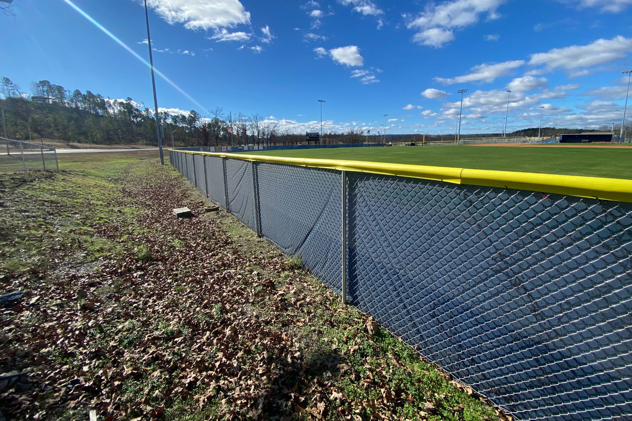 A sunny outdoor baseball or softball field with a yellow-topped chain-link fence, green grass, scattered leaves on the ground, and a clear blue sky with white clouds.