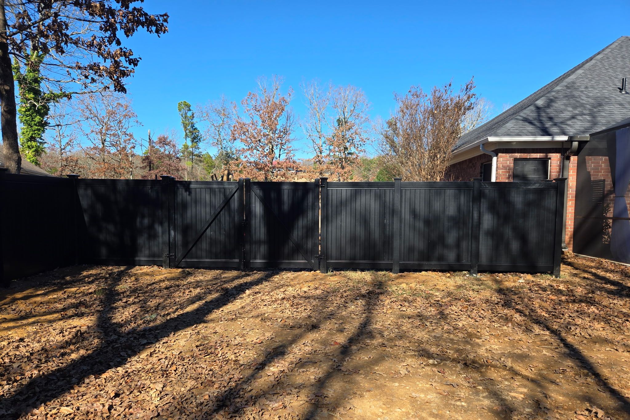A backyard with a black privacy fence, bare leaf-covered ground, and some trees with autumn foliage; a brick house is partially visible on the right side under a clear blue sky.