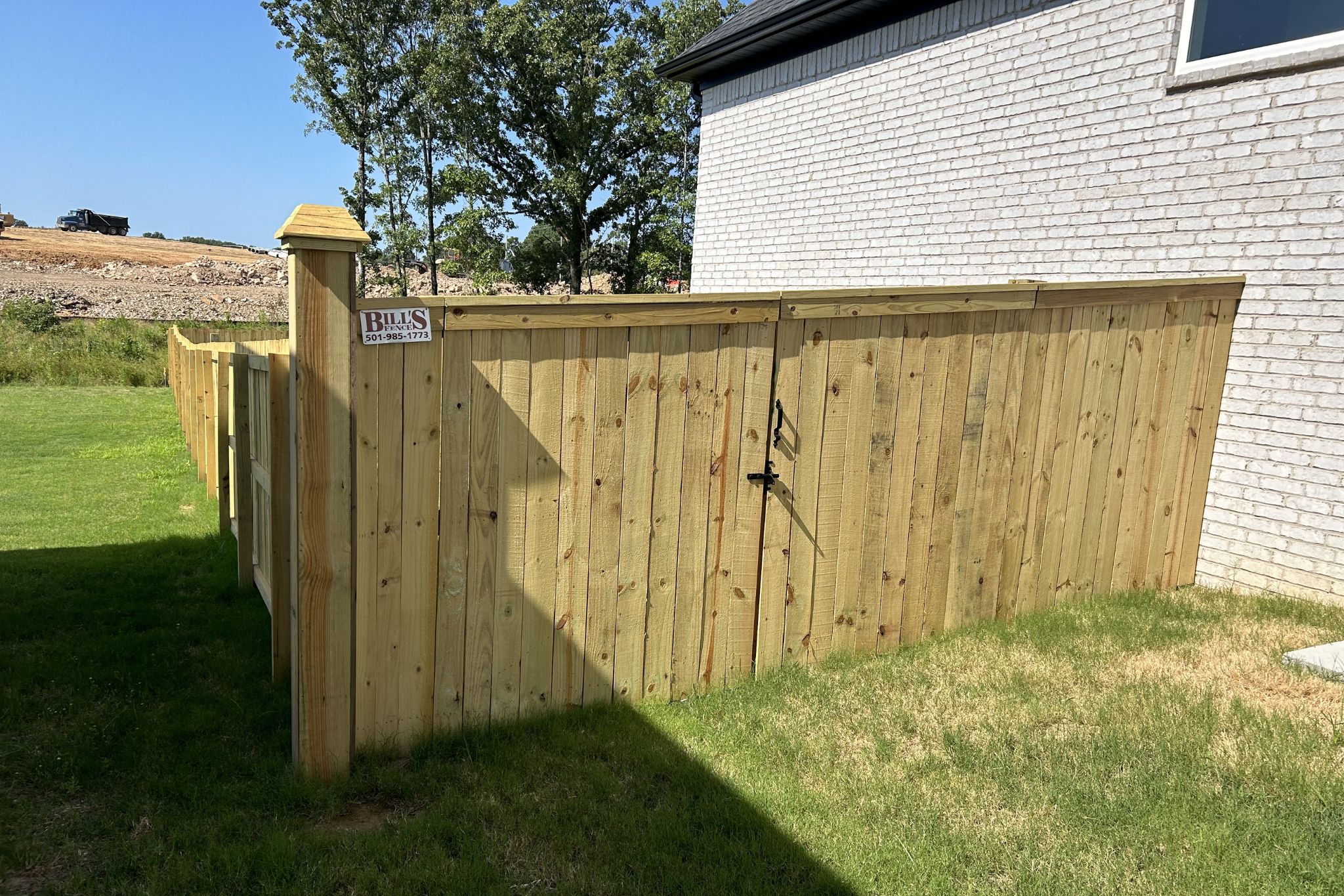 A tall wooden privacy fence encloses a grassy yard next to a white brick house. The fence has a locked gate and a sign reading Bills Construction. Trees and a construction site are visible in the background.
