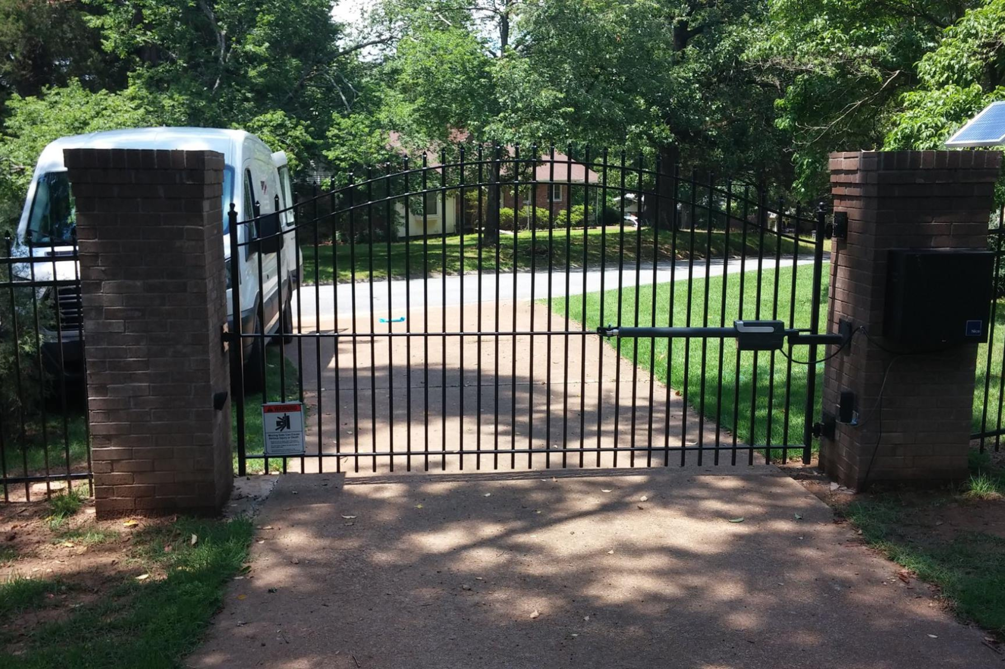 A black metal driveway gate with brick pillars blocks the entrance to a residential property; a white van is parked on the left, and trees and grass surround the area.