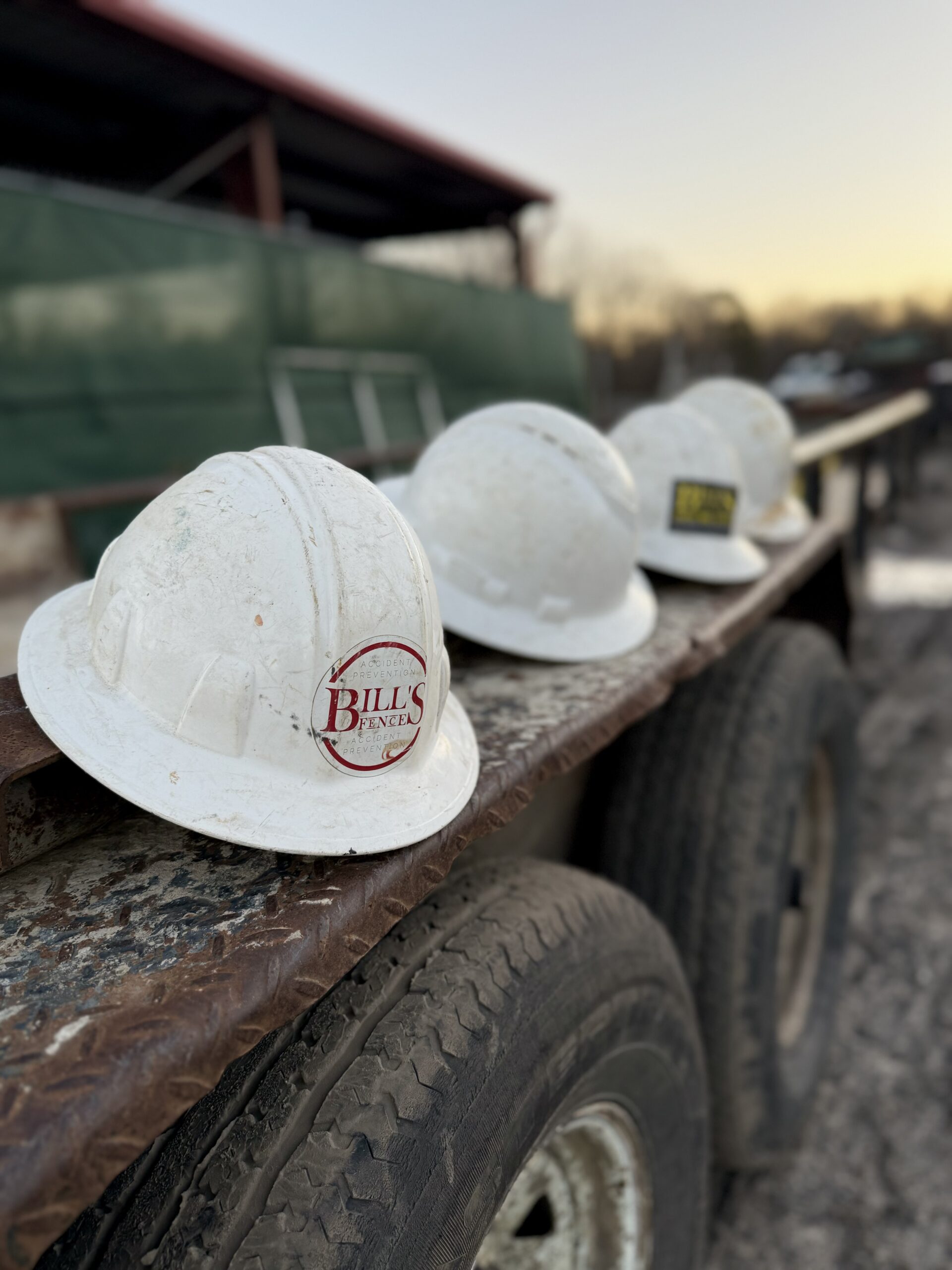 Four white construction hard hats, one labeled “Bill’s Fence,” rest in a row on the rusty edge of a utility trailer with worn tires. A building and fencing are blurred in the background at sunset.