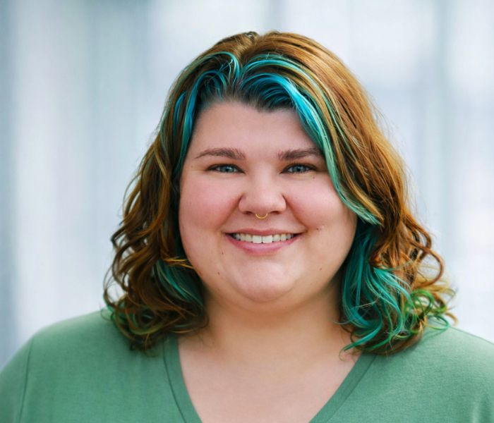 A person with wavy, shoulder-length brown hair featuring teal highlights smiles at the camera. They are wearing a green top and have a septum piercing. The background is softly blurred.