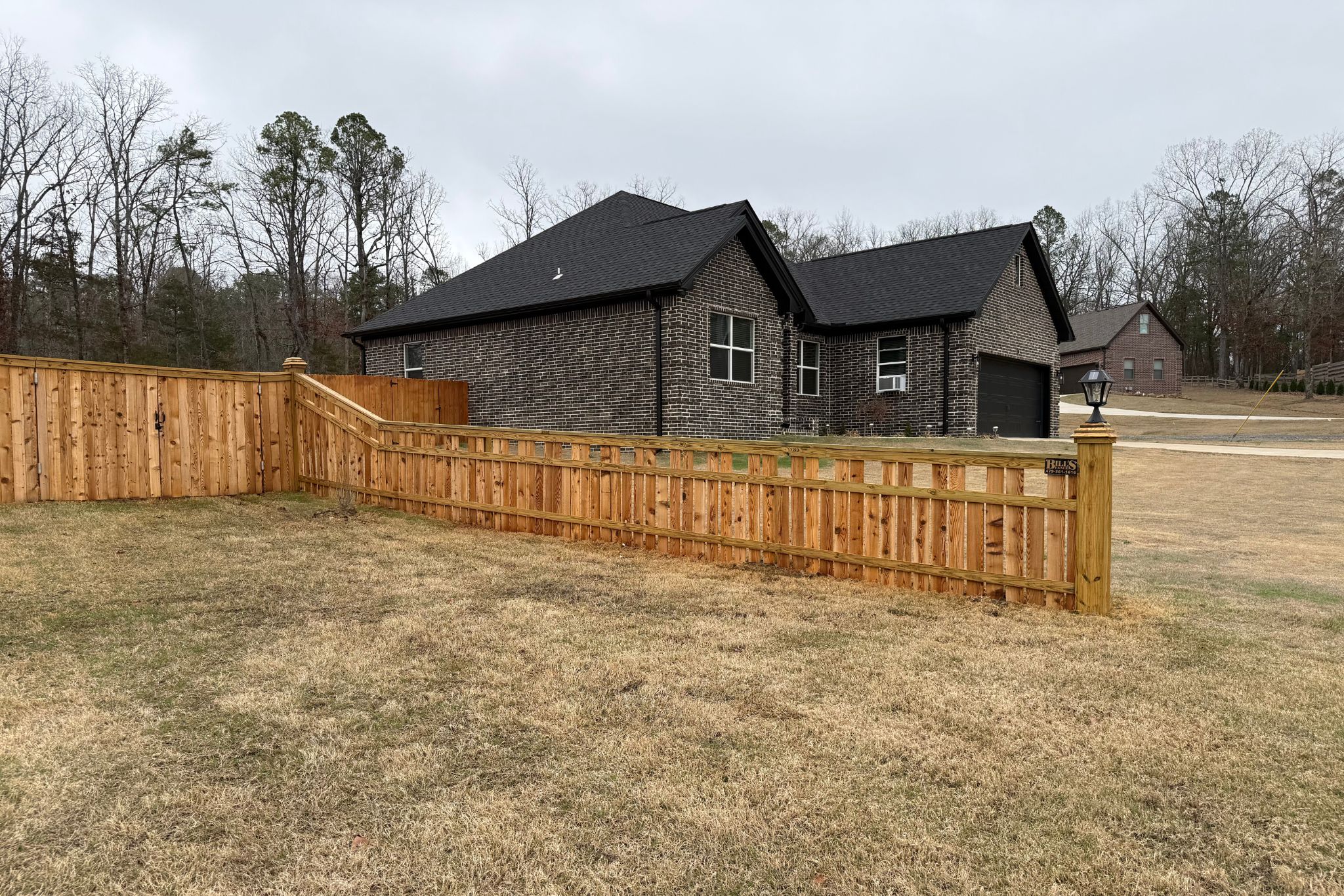 A single-story brick house with black roof stands behind a wooden fence on a grassy yard. Leafless trees and another house are visible in the background under a cloudy sky.