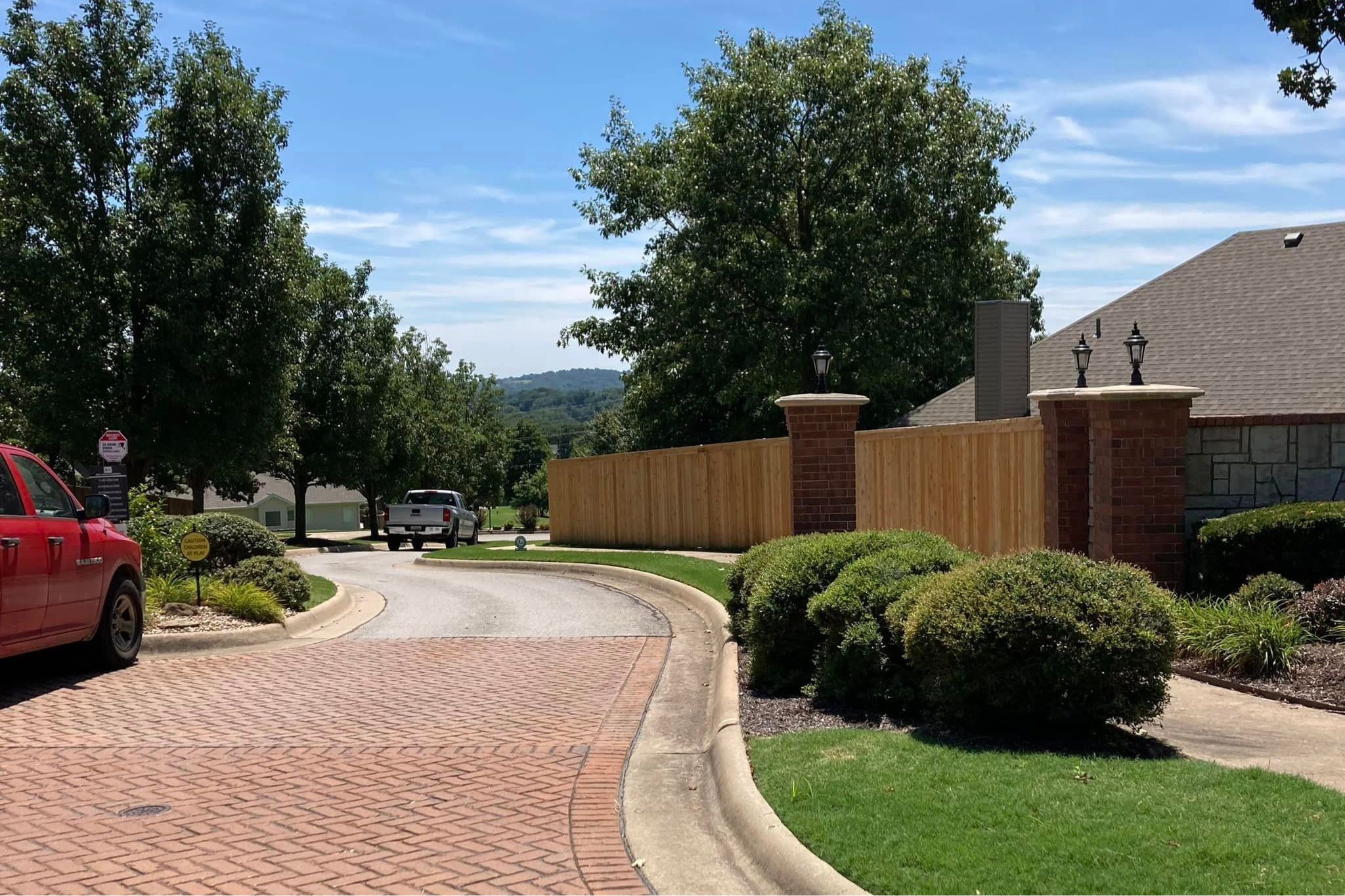 A suburban street with brick pavement curves past manicured bushes, a wooden fence, and brick pillars. A white truck and red vehicle are parked near green trees under a blue sky.