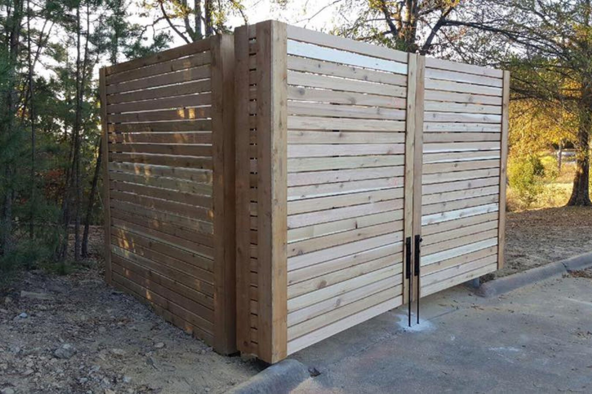 A wooden enclosure with vertical and horizontal slats stands on a concrete pad next to a paved area, surrounded by trees and natural ground cover. The enclosure has a double-door gate with black handles.