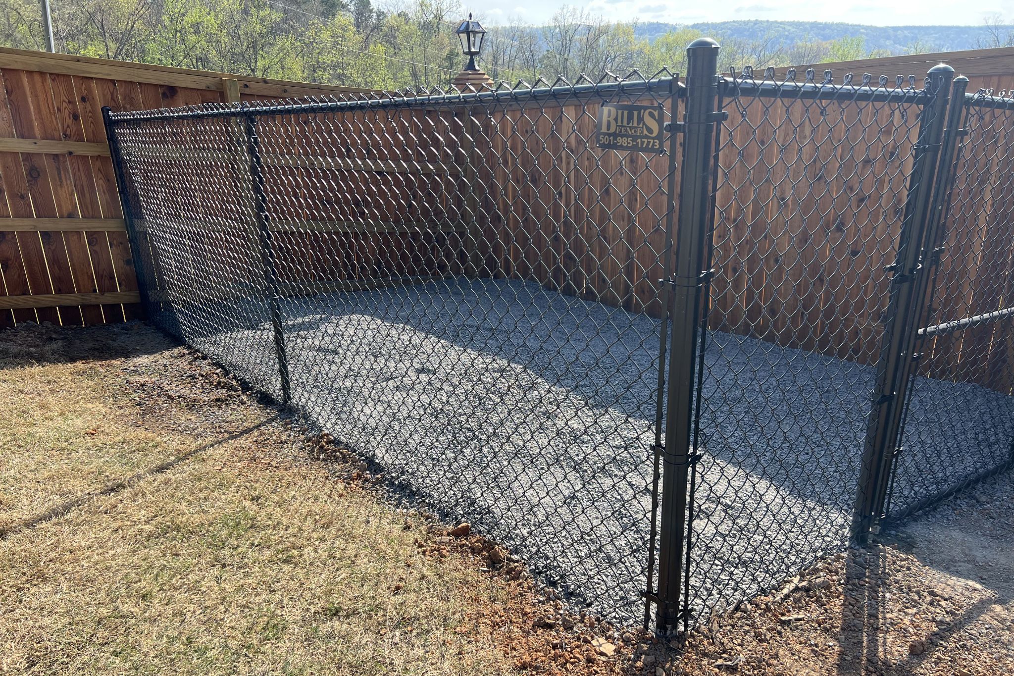 A fenced outdoor enclosure with a black chain-link fence set on gravel, bordered by wooden fencing and a grassy lawn, under a sunny sky.