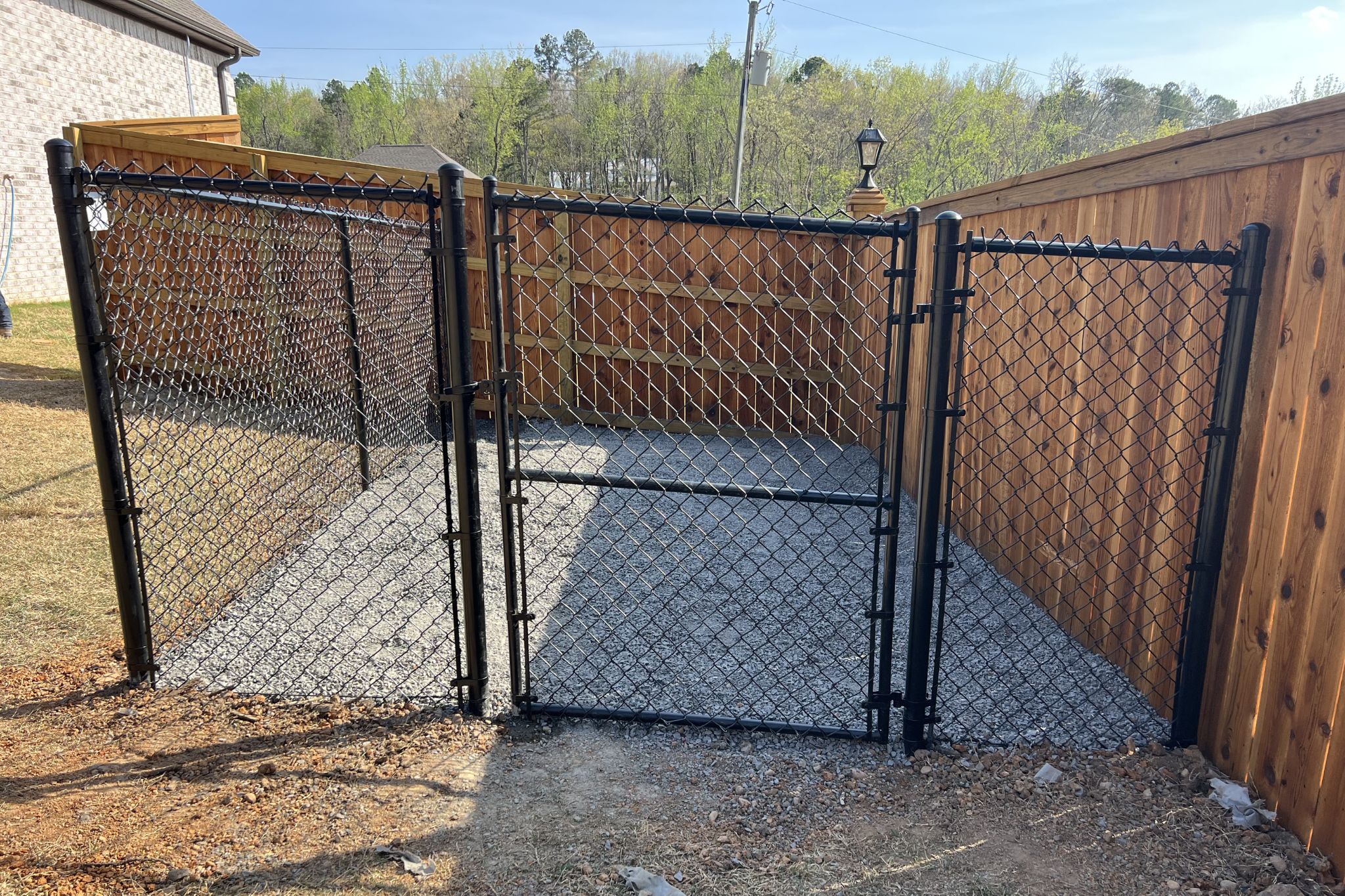 A fenced outdoor enclosure with black chain-link gates and wooden privacy fences on two sides, set on a gravel surface with trees and a house visible in the background.