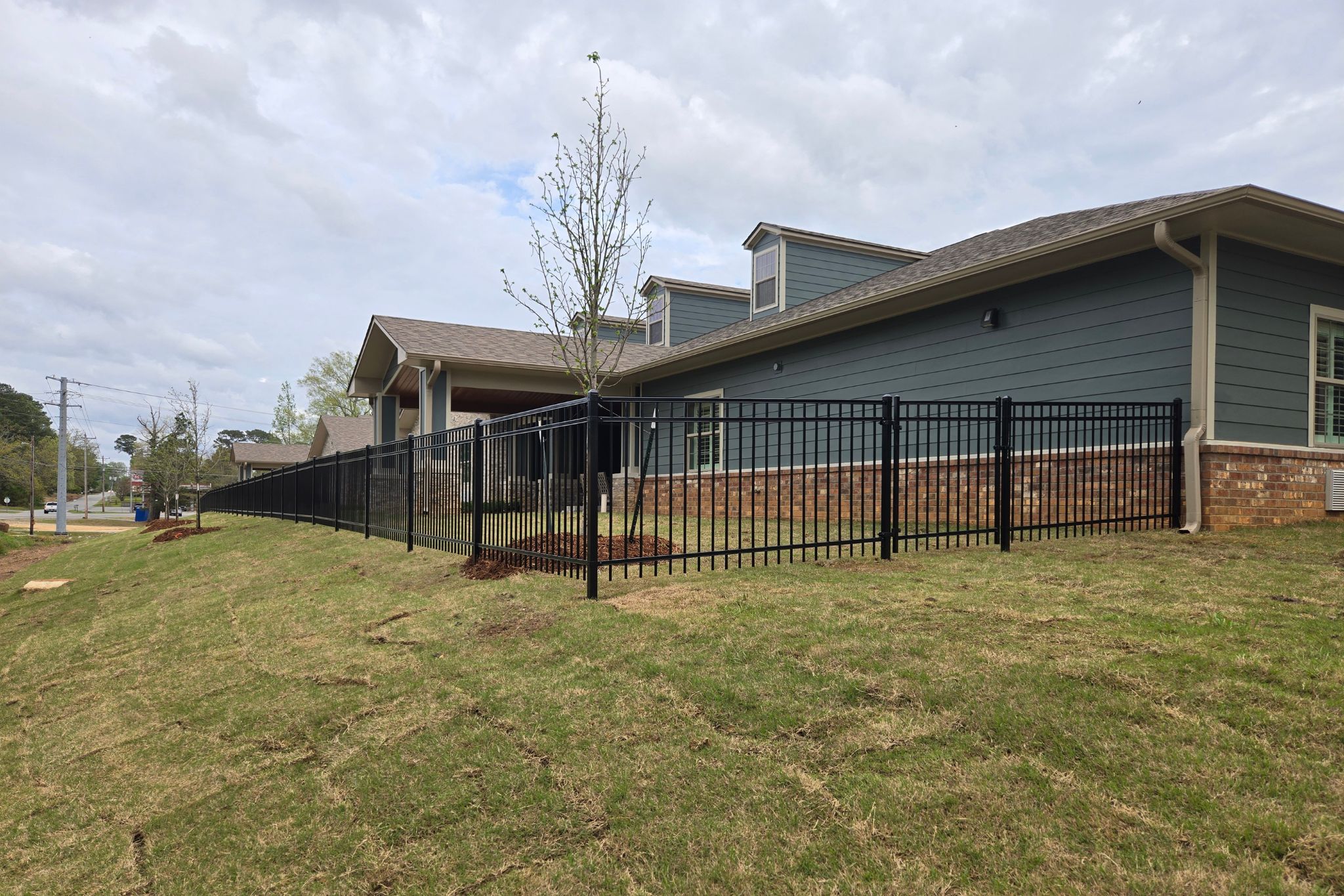 A modern building with gray siding and brick accents sits behind a black metal fence on a grassy, sloped lawn. Young trees are planted along the fence, and the sky is mostly cloudy.