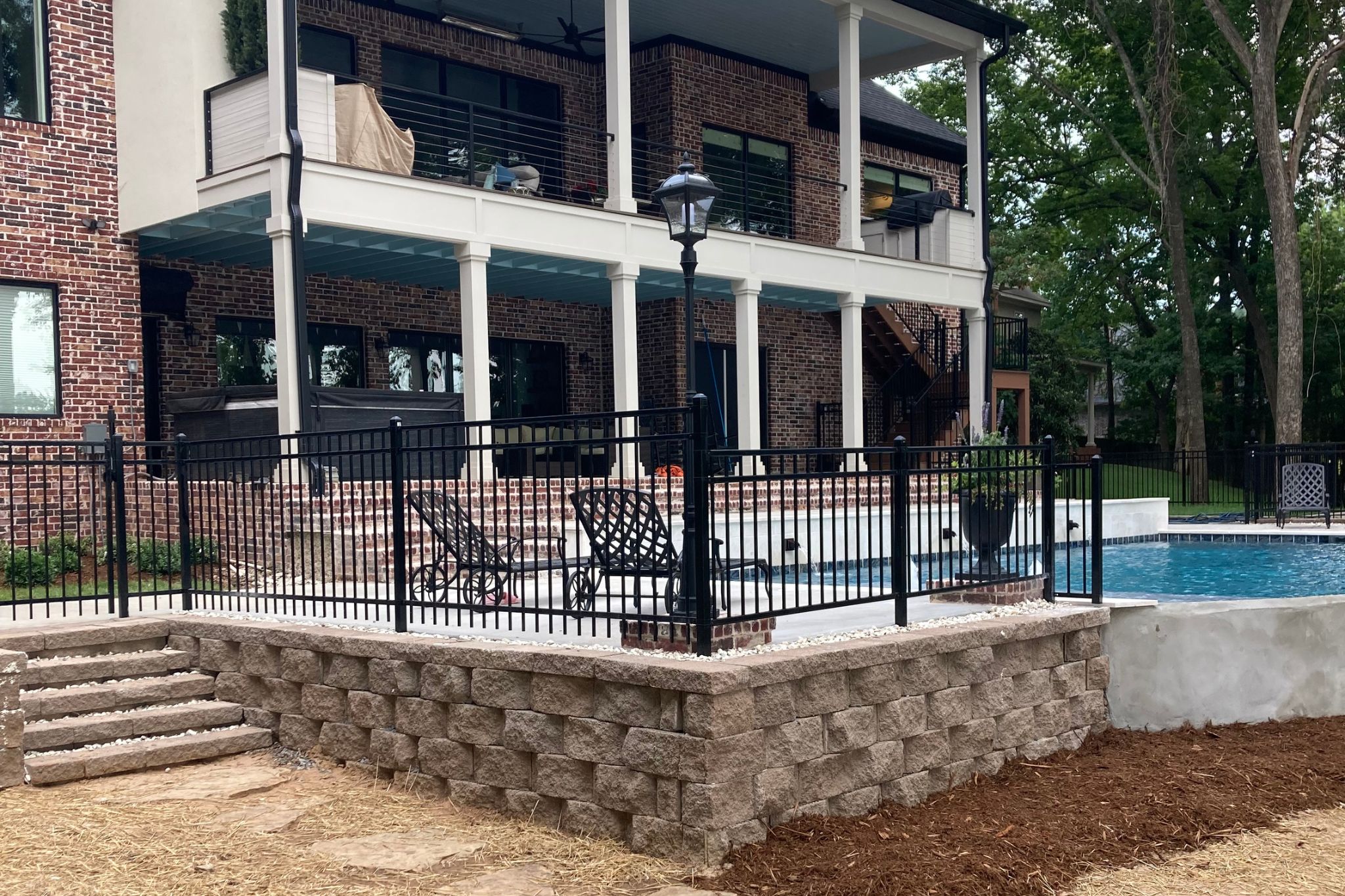 A backyard with a stone retaining wall, black metal fence, in-ground pool, lounge chairs, and a two-story brick house with covered patios and outdoor seating areas. Trees and greenery surround the yard.