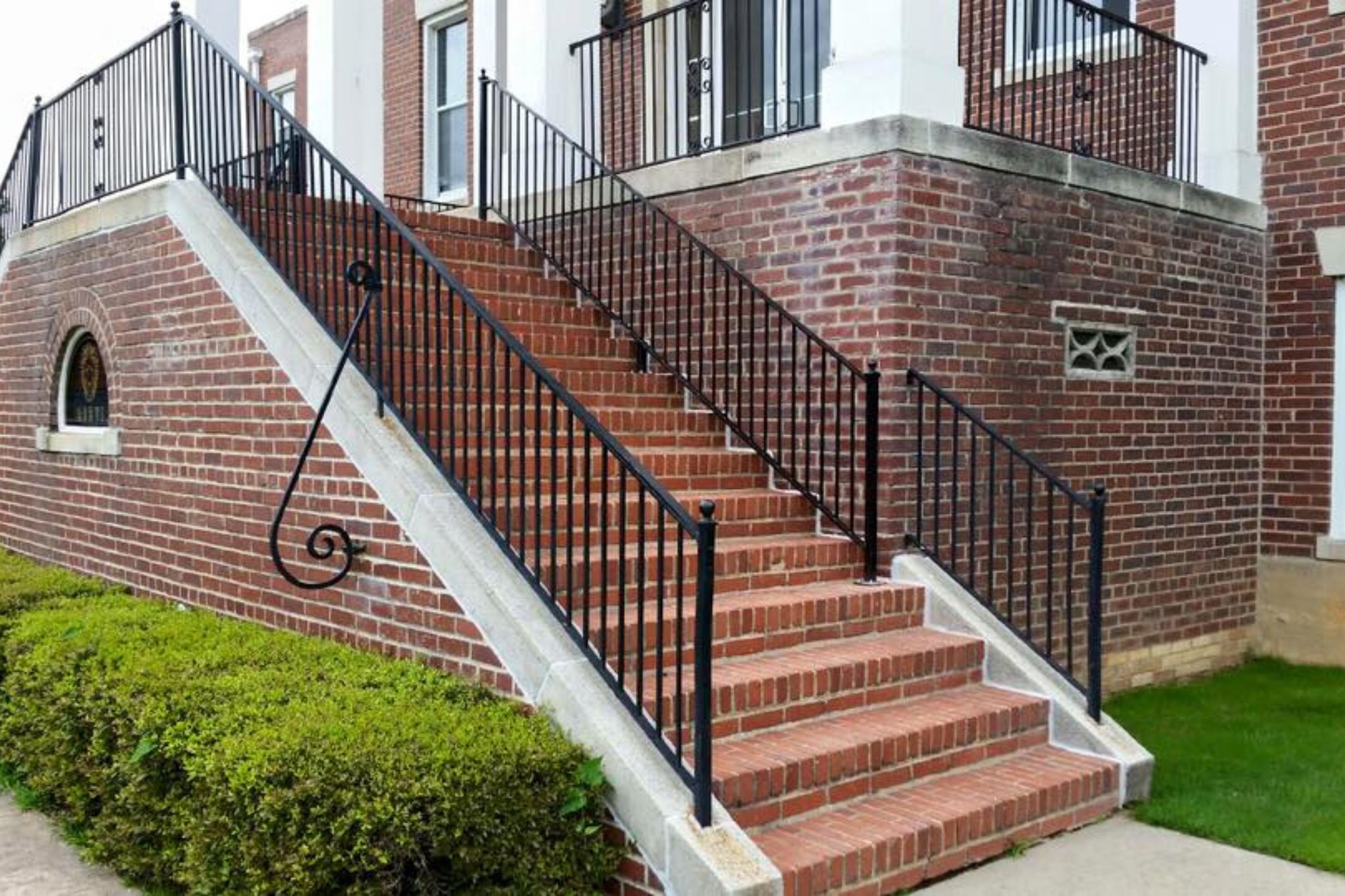 A brick staircase with black metal railings leads up to the entrance of a brick building. There is a green shrub along the base of the stairs and a small rounded window in the brick wall.