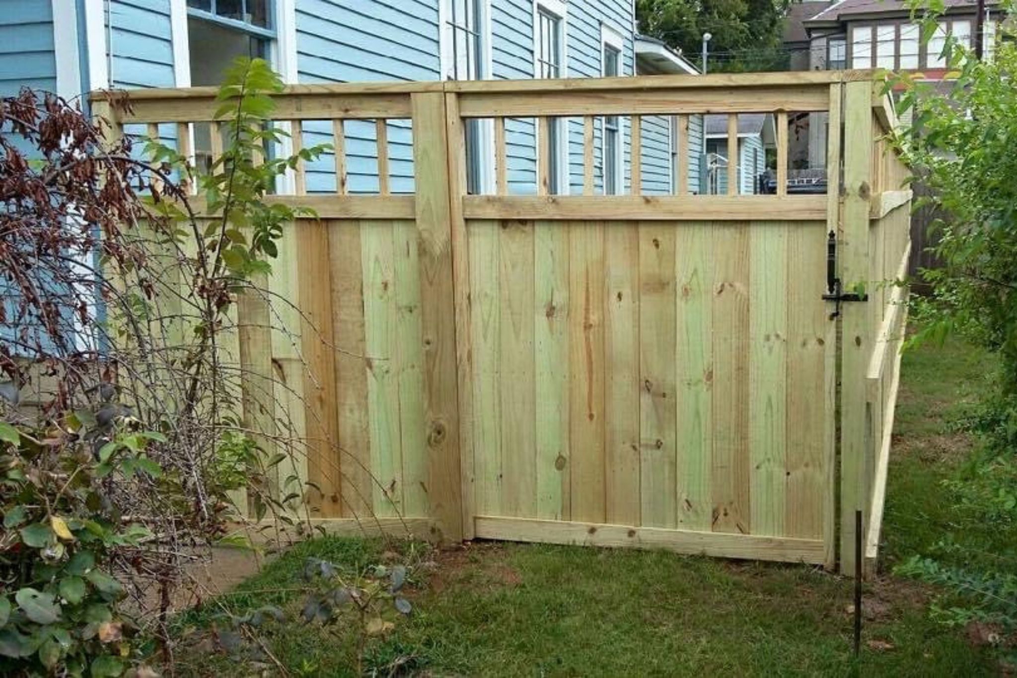 A newly built wooden fence with a gate stands next to a blue house. The fence has vertical boards and decorative lattice panels on top. Green grass and some plants grow along the fence.
