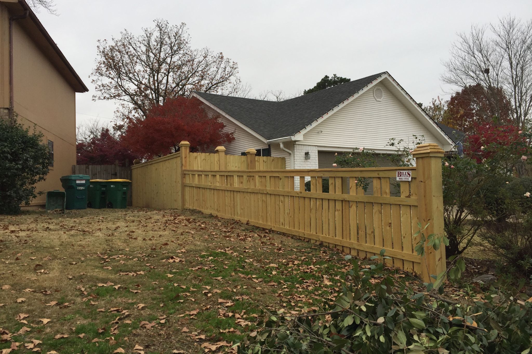 A newly built wooden fence runs alongside a grassy yard with scattered leaves. Green trash bins line the fence near a beige house, and a white house with a dark roof is visible in the background.