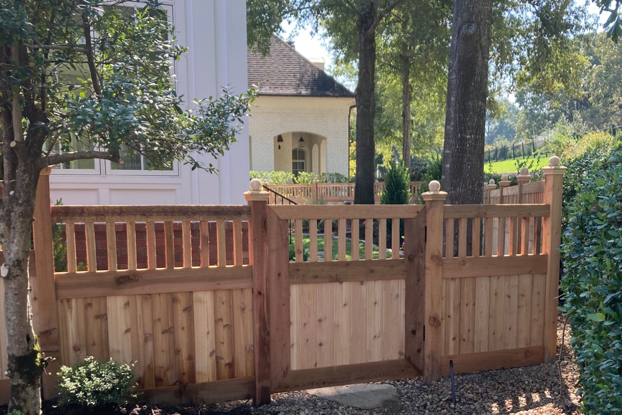 A tall wooden privacy fence encloses a grassy yard next to a white brick house. The fence has a locked gate and a sign reading Bills Construction. Trees and a construction site are visible in the background.