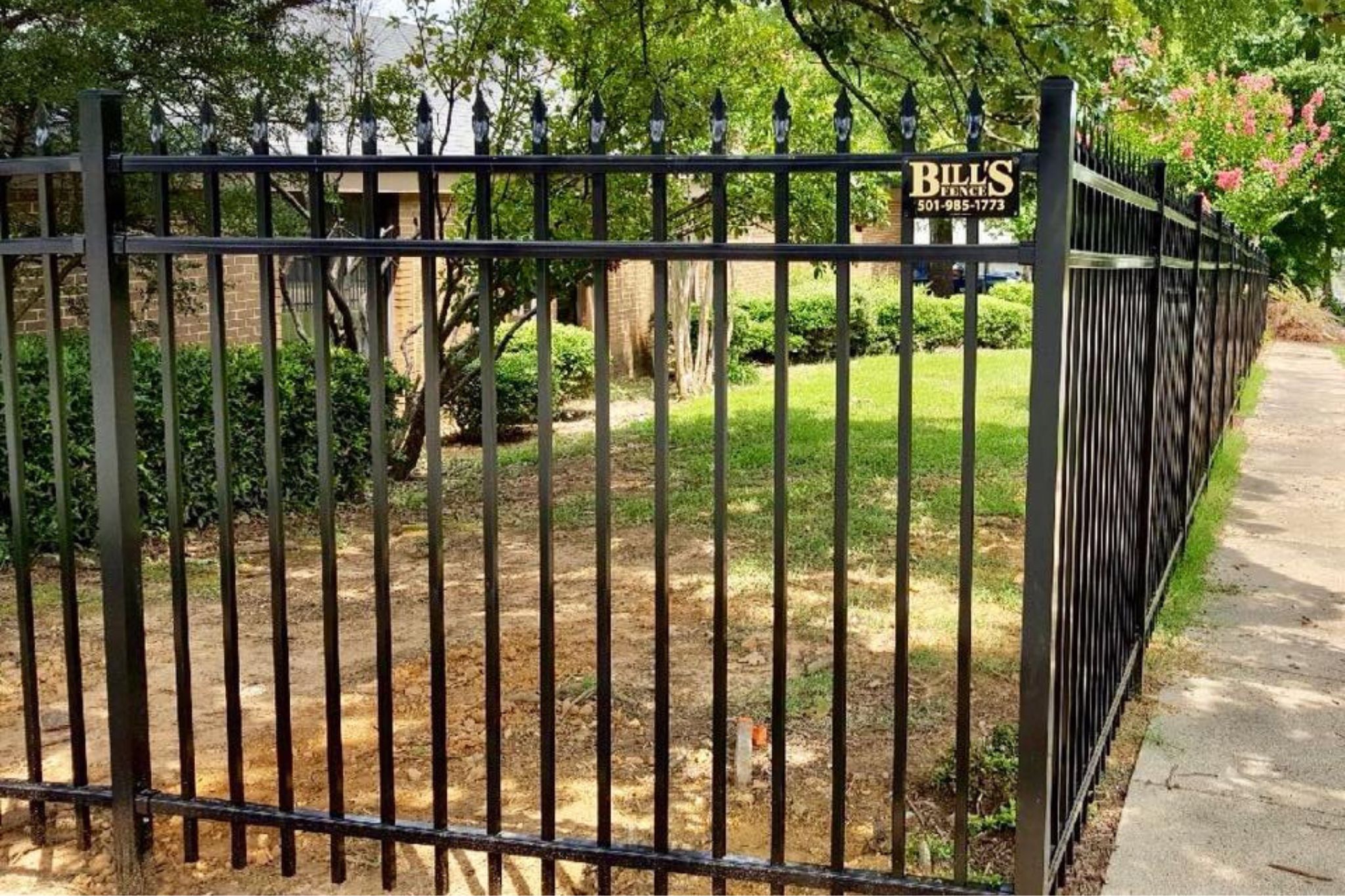 Black metal fence with pointed tops surrounds a grassy, landscaped yard with bushes and trees. A sign reading Bills Fence Co. 501-985-1773 is attached to the fence. Sidewalk and street visible on the right.
