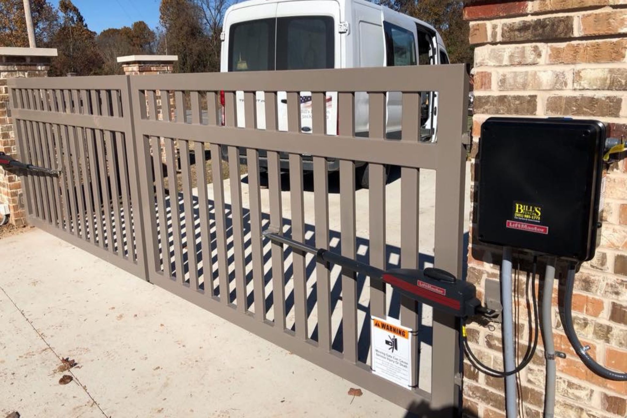 A beige automatic metal gate with safety and control boxes attached, partially open across a driveway. A white van is parked behind the gate, and brick pillars frame the entrance.