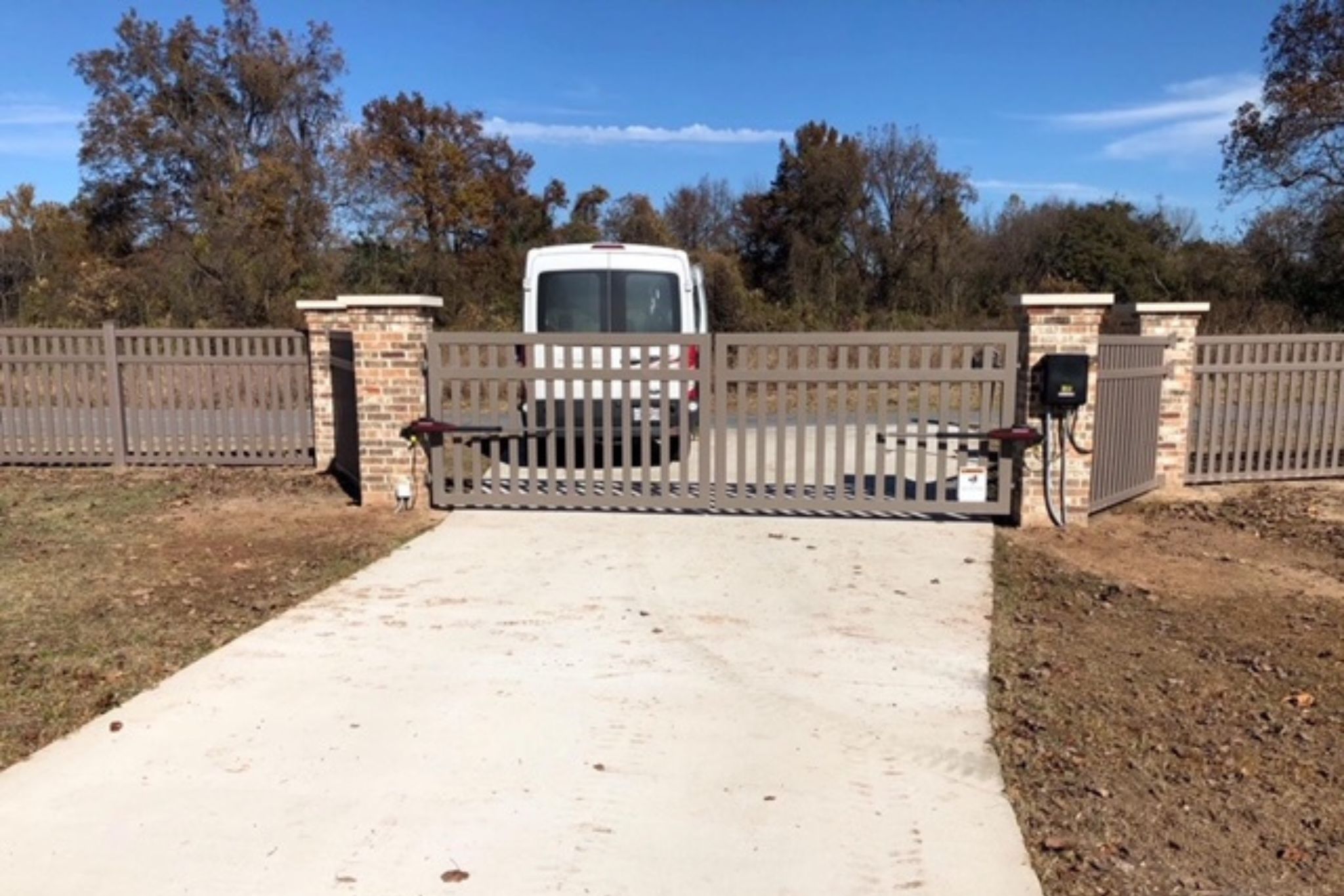 A white van is parked behind a closed, automated metal gate with brick pillars at the entrance of a fenced property. The driveway is concrete and trees are visible in the background under a blue sky.