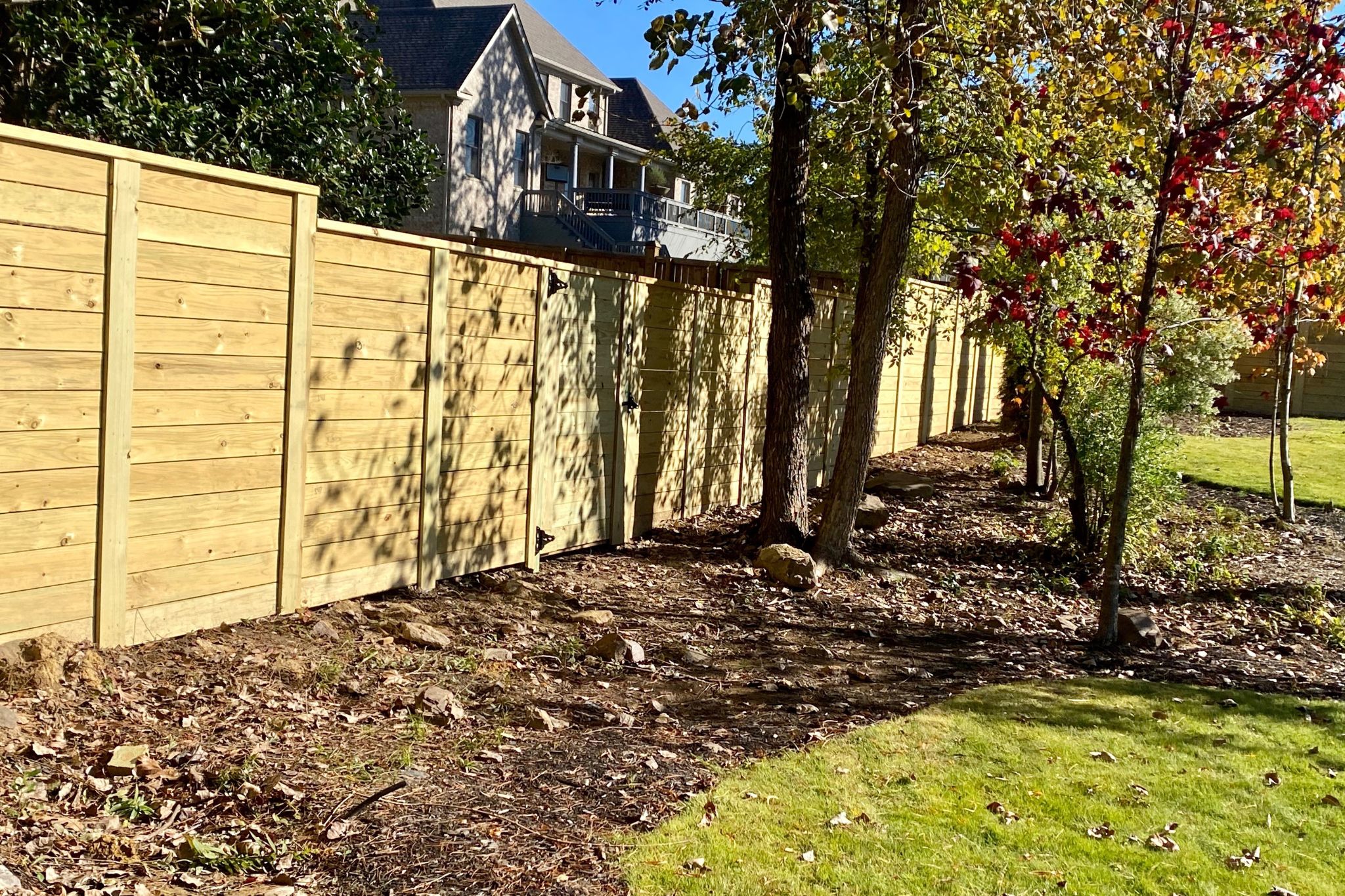 A tall wooden privacy fence runs along the edge of a yard with grass, trees, and patches of dirt. A house with a balcony is visible in the background under a clear, sunny sky.
