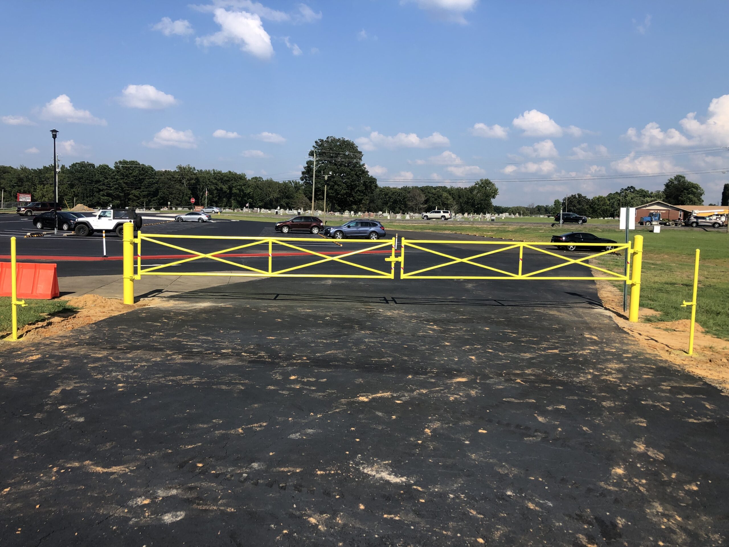 A bright yellow metal gate and fence block the entrance to a paved road, with cars and a parking lot visible in the background under a partly cloudy blue sky.