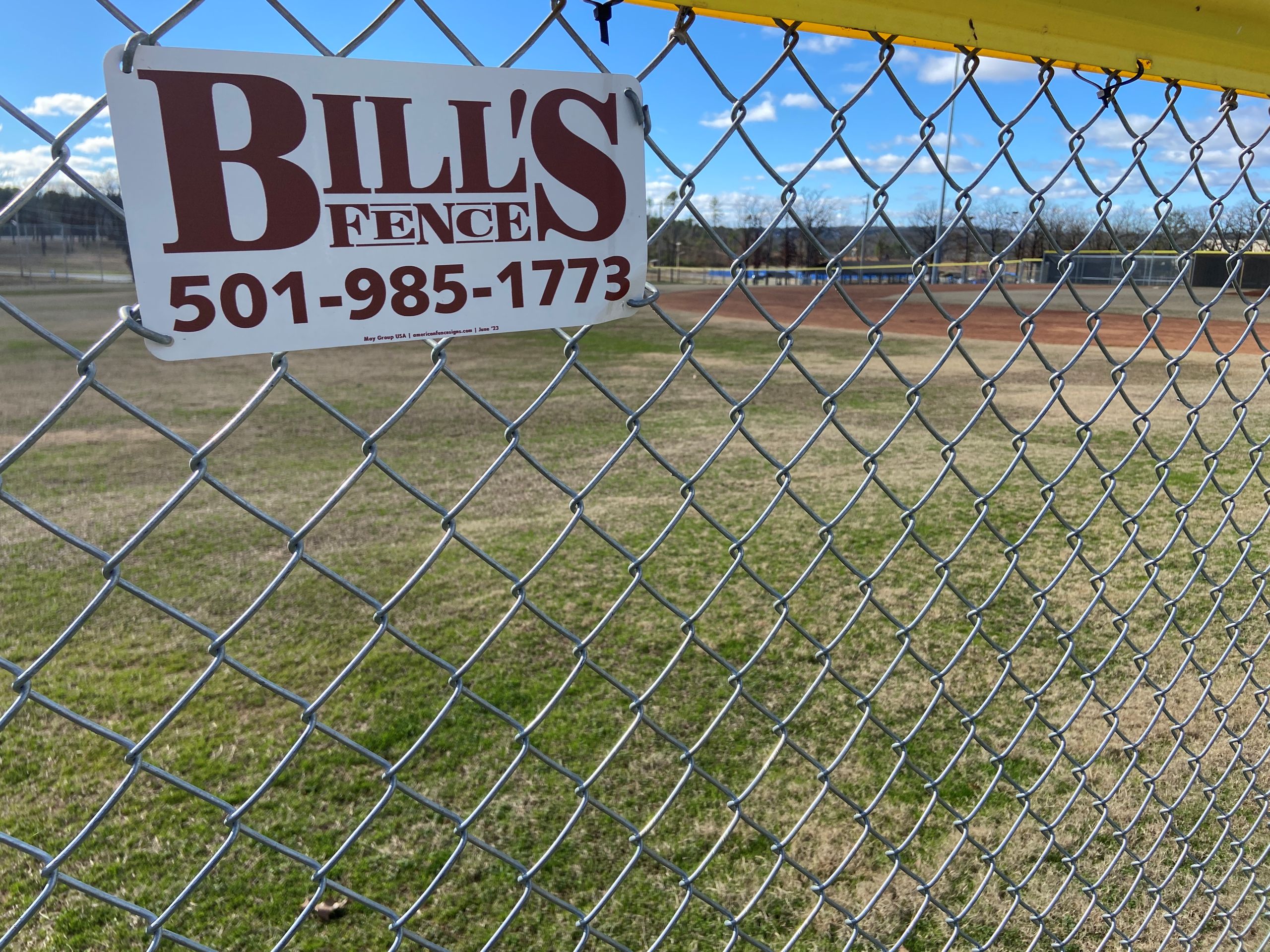 A sign reading BILLS FENCE 501-985-1773 is attached to a sturdy chain-link fence, with a grassy field and a baseball diamond stretching into the background beneath a clear blue sky.