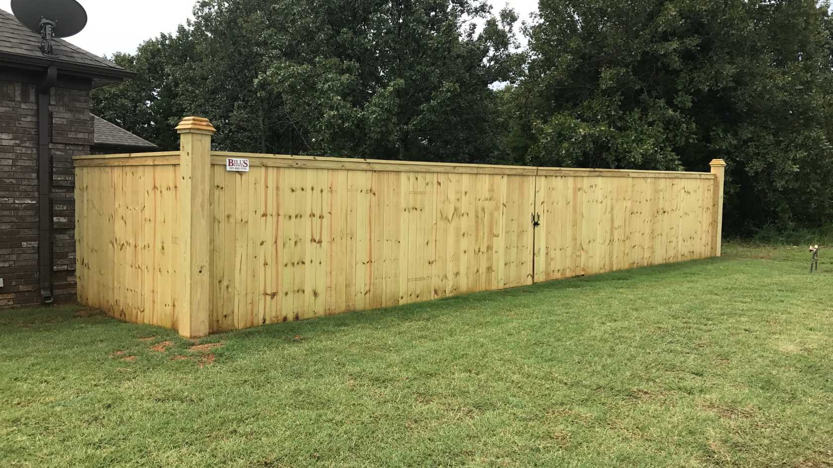 A tall wooden privacy fence with a latch, set in a grassy yard beside a brick building, with green trees in the background.