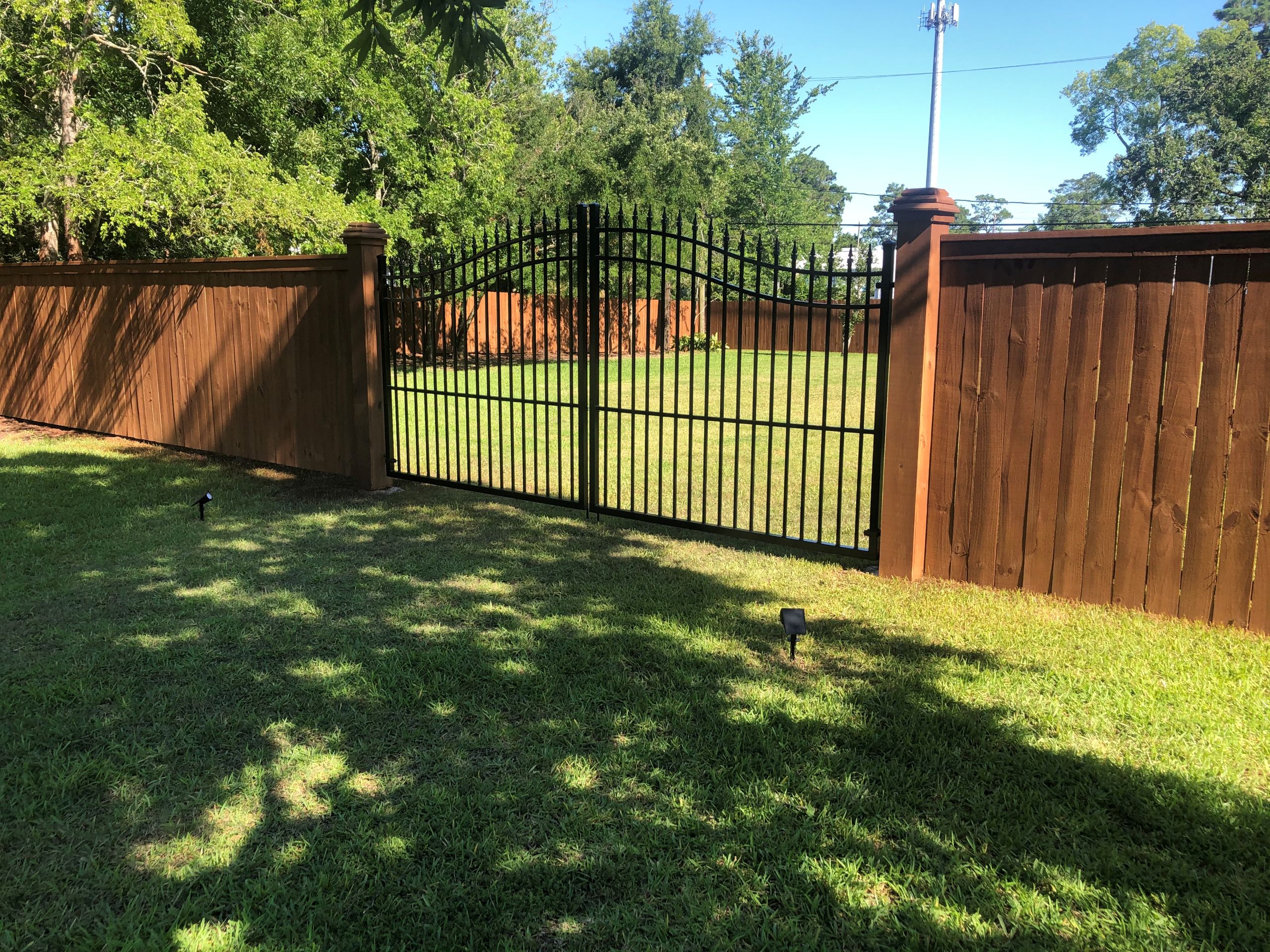 A black metal gate by Bill's Fence Co is set in a tall wooden fence, opening onto a grassy yard with trees and sunlight casting shadows on the lawn.